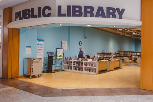Wide entrance to a room with books and a smiling person at a service desk. Public Library in written above the entrance.
