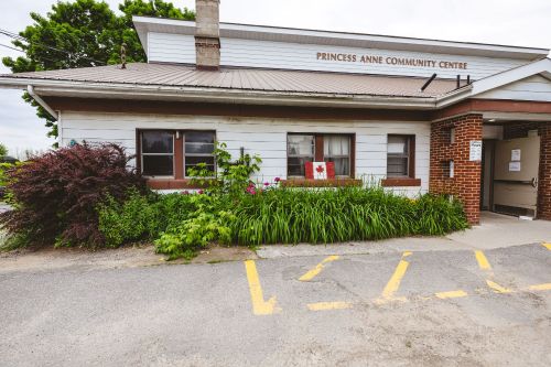 Small house with white siding, green bushes, Canadian flag and small sign for hours of operations