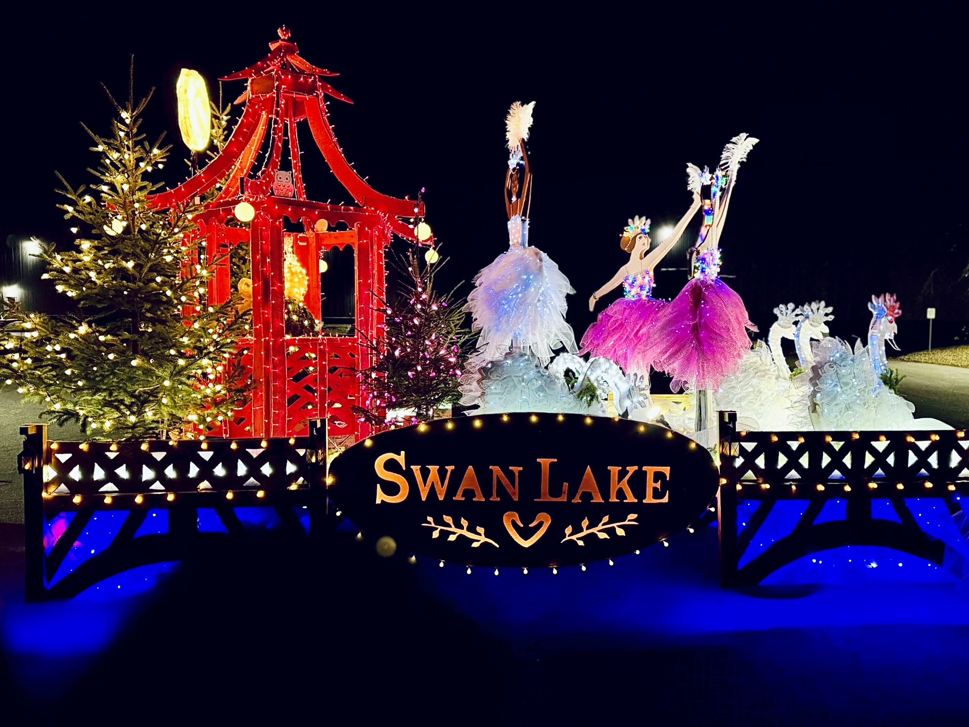 An image of the KFPL float. The theme is Swan Lake. There are wooden cutouts of ballet dancers, a pavilion in red lights, white swans, and christmas trees on the float. There is a sign that reads Swan Lake.