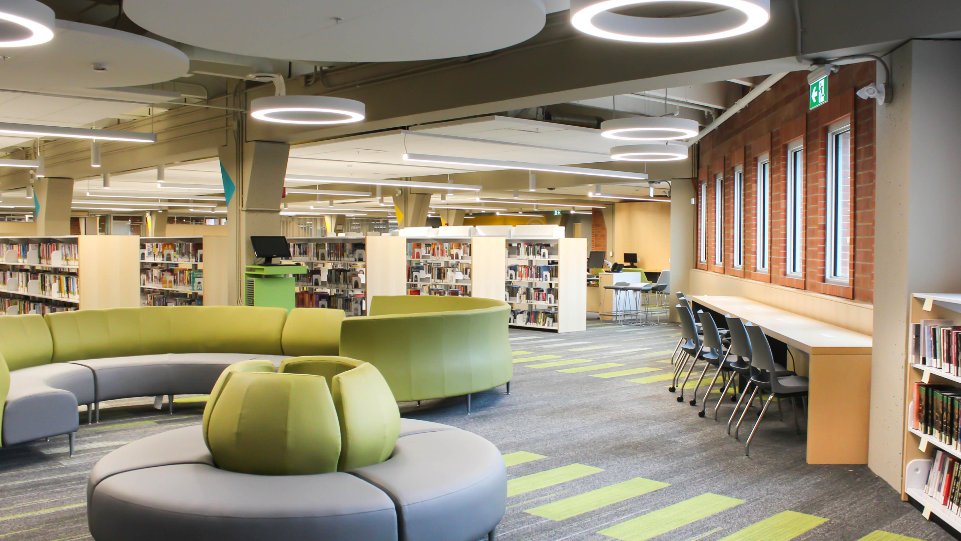 The interior of the Central Branch, showing curved grey and green seating, light wood bookshelves, circular overhead lights, and a row of chairs against a light wood desk below a window.